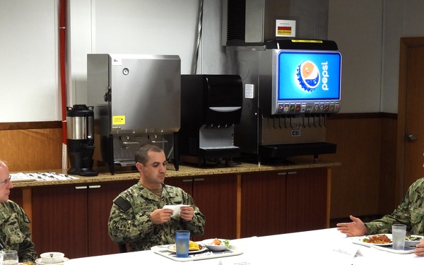 Three military personnel in U.S. Navy camouflage uniforms (NWU Type III) are seated at white-tablecloth-covered tables in what appears to be a military dining facility (DFAC/mess hall). They appear to be having a working lunch or formal dining meeting, with food plates and beverages visible on the tables. A Pepsi fountain dispenser and coffee equipment are visible in the background.
