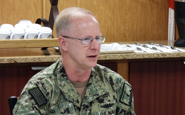 A U.S. Navy officer identified by his name tape as 'Caudle' is seated at a dining table, apparently speaking during a meal or briefing event. A place card in front of him reads 'Vice Admiral Daryl Caudle, Commander, Submarine Forces.' He is wearing Navy Working Uniform (NWU) digital camouflage with U.S. flag patch and submarine warfare insignia.