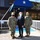 Three individuals pose for a formal photograph outside the Commander, Undersea Warfighting Development Center building. Two are wearing U.S. Navy camouflage uniforms with face masks, flanking a civilian in a dark suit also wearing a face mask. The setting appears to be an official military installation visit or ceremony.