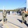 A group of U.S. Navy personnel in camouflage uniforms and face masks are exchanging salutes on a naval pier or waterfront facility. The scene appears to be a formal military greeting or ceremony conducted outdoors at what looks like a naval base. Industrial crane equipment and waterfront infrastructure are visible in the background.