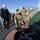 A civilian official in a dark suit is being given a tour or briefing on what appears to be the deck of a submarine by U.S. Navy personnel in camouflage uniforms. Several sailors are crouching near an open hatch on the submarine's hull, appearing to explain or demonstrate its features. All individuals are wearing face masks, suggesting this was taken during the COVID-19 pandemic period.