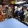 Four individuals wearing face masks are gathered inside what appears to be the wardroom or briefing area of USS Colorado (SSN-788), a U.S. Navy submarine. A Navy sailor in camouflage uniform is gesturing toward a plaque or display on the wall, appearing to brief two civilians and a senior Navy officer. The setting suggests an official tour or visit to the submarine.