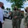 Four individuals are gathered outdoors in a parking lot area, with three wearing U.S. Navy camouflage uniforms and one in civilian business attire. All four are wearing black face masks, suggesting this was taken during the COVID-19 pandemic period. The group appears to be engaged in a briefing or discussion, likely at a naval installation based on the visible vehicle markings.