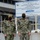 Two U.S. Navy personnel in camouflage uniforms and black face masks are standing in front of a project sign at Naval Submarine Base New London in Groton, CT. The sign references an Energy Savings Performance Contract (Contract No. N39430-20-F-9905) valued at $83M, dated December 2019, managed by NAVFAC Mid-Atlantic and contractor NORESCO of Westborough, MA. The individuals are identified by name tags as 'CAUDLE' (left, wearing a gold cap) and 'MOORE' (right, wearing a tan cap).