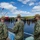 Three U.S. Navy personnel in digital camouflage uniforms (NWU Type III) stand on a pier or dock, observing waterfront construction or dredging operations. They are viewed from behind, looking out over a waterway with industrial equipment including a large crane barge and construction infrastructure. One individual on the right is wearing a face mask.