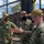 Three U.S. Navy personnel in digital camouflage uniforms (NWU Type III) are engaged in a discussion outdoors at what appears to be a military installation. All three are wearing black face masks and tan/coyote brown baseball caps, consistent with COVID-era protocols. One individual appears to be gesturing with both hands, suggesting an active briefing or explanation.