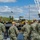 A group of approximately six military personnel in U.S. Navy NWU Type III (digital camouflage) uniforms and tan caps observe waterfront industrial operations from a dock or pier. They are watching a large crane barge numbered '526' and associated heavy equipment at what appears to be a naval or industrial waterfront facility. Several personnel are wearing face masks, suggesting this was taken during the COVID-19 pandemic period.