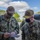 Two U.S. Navy personnel in NWU Type III digital camouflage uniforms are reviewing a document together outdoors. The individual on the left is identified by name tape as 'KOERNKE' and the one on the right as 'CAUDLE', who displays three-star rank insignia and wears a 'COMSUBFOR' (Commander, Submarine Forces) cap. Both are wearing black face masks, suggesting this was taken during the COVID-19 pandemic period.