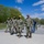 A group of military personnel in U.S. Navy Working Uniform (NWU Type III) camouflage are walking along a paved path at what appears to be a military installation or base. All uniformed individuals are wearing black face masks and tan/coyote brown baseball caps. A civilian in casual clothing is visible in the background near what appears to be a flagpole area.
