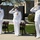 Submarine Squadron Four welcomes new commodore Three U.S. Navy officers in white dress uniforms are standing at salute during an outdoor military ceremony. They are wearing face masks and display multiple service medals and ribbons on their uniforms. A dark memorial wall with engraved names is visible in the background.