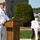 Submarine Squadron Four welcomes new commodore Two U.S. Navy officers in white dress uniforms are participating in an outdoor military ceremony, likely a change of command or memorial event. One officer stands at a wooden podium speaking into a microphone, while a second officer wearing a face mask is seated behind him. The setting appears to be a naval installation with a waterfront visible in the background.