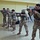 Five military personnel in camouflage uniforms are practicing rifle aiming stances inside an indoor facility, likely a training room or gymnasium. They are lined up in a staggered formation, all shouldering what appear to be M16 or M4-style rifles with slings attached. This appears to be an indoor weapons handling or marksmanship training exercise.