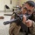 A U.S. Army soldier in MultiCam/OCP camouflage uniform is lying in a prone position on an indoor floor, aiming a rifle (appears to be an M16-style weapon) toward the camera. The setting appears to be a cafeteria or multipurpose room, suggesting a training exercise or demonstration. The soldier wears glasses and has a U.S. flag patch and a unit patch visible on his uniform.