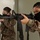 Two female U.S. Army soldiers in Operational Camouflage Pattern (OCP) uniforms are conducting weapons training, aiming M16-style rifles in an indoor environment. Both wear black face masks and display unit patches on their left shoulders. The image appears to be a training exercise, likely in a military facility.