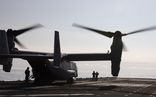 A tiltrotor aircraft, identifiable as a Bell Boeing V-22 Osprey, is positioned on the flight deck of a naval vessel with its rotors spinning. The image is taken from a low angle looking along the deck toward the horizon, with several personnel visible around the aircraft. The strong backlighting creates a silhouette effect against an overcast sky and open ocean.