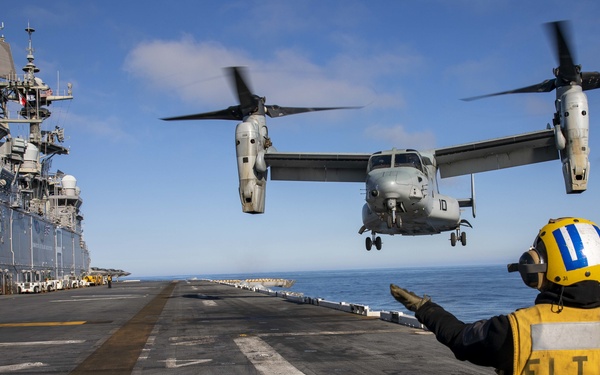 A flight deck crew member wearing a yellow 'FLT DK' vest and yellow helmet is directing an MV-22 Osprey tiltrotor aircraft during a landing or takeoff operation on a naval vessel's flight deck. The ship's island superstructure is visible on the port side, and the operation is taking place at sea under clear blue skies. The rotors of the Osprey are in motion, indicating active flight operations.