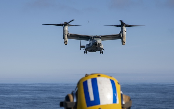 A military flight deck crew member in yellow safety gear and helmet guides a V-22 Osprey tiltrotor aircraft approaching a naval vessel for landing. The scene takes place on a ship's flight deck over open ocean under clear skies. The Osprey's rotors are in the vertical/helicopter configuration, indicating it is in hover or landing mode.