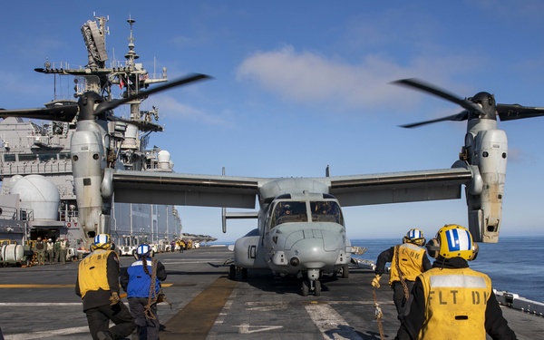 Flight deck crew members in yellow vests and helmets are working around a V-22 Osprey tiltrotor aircraft on the deck of a naval amphibious assault ship. The rotors are spinning, indicating the aircraft is either preparing for takeoff or has just landed. Additional naval vessels are visible in the background on open ocean.