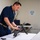 A military or naval personnel member in dark blue coveralls is handling and inspecting a machine gun or light weapon on a white table in what appears to be an interior shipboard or facility room. The individual appears to be conducting weapons maintenance, inspection, or instruction. A sign on the wall reads '4-35-2-Q / FR 35-42 / EXEC', suggesting a naval vessel compartment designation.