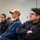 A group of individuals, several wearing blue military-style uniforms, are seated and attentively watching something off-camera in what appears to be a briefing or classroom setting. The foreground subject, wearing glasses and a black jacket with arms crossed, appears focused and engaged. The setting suggests a formal training or instructional session.