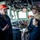 Three U.S. Navy personnel are engaged in conversation on the bridge of a naval vessel. The individual on the left, wearing an orange cap and dark coveralls with visible U.S. Navy insignia, appears to be briefing or discussing something with two other crew members wearing olive/tan caps. The setting is a ship's bridge with navigation equipment, controls, and instrumentation visible in the background.