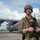 75th EAS Executes Tactical Airlift Mission in Manda Bay, Kenya A military service member in full combat gear stands on an airfield tarmac in front of a large gray military transport aircraft. The soldier is equipped with a tactical helmet, communications headset, body armor, and is holding an assault rifle. The setting appears to be an outdoor airfield under partly cloudy skies.