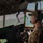 75th EAS Executes Tactical Airlift Mission in Manda Bay, Kenya A military aviator in camouflage uniform and Bose headset is seated in the cockpit of a large military transport aircraft, likely a C-130 Hercules, flying over a coastal or oceanic area. The cockpit features extensive avionics, multi-function displays, and a heads-up display (HUD) mounted on the instrument panel. The pilot appears to be actively monitoring the flight, with water and coastline visible through the windscreen.