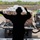 75th EAS Executes Tactical Airlift Mission in Manda Bay, Kenya A military loadmaster or ground crew member, silhouetted against bright daylight, signals with raised arms to guide a cargo vehicle backing into what appears to be the rear ramp of a military transport aircraft. The vehicle is loaded with multiple palletized and strapped cargo bundles. The scene takes place on a military installation or airfield, with concrete barriers and green vegetation visible in the background.