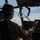 75th EAS Executes Tactical Airlift Mission in Manda Bay, Kenya A military aviator in camouflage uniform is seated in the cockpit of a military aircraft, appearing to operate or gesture toward the instrument panel. The pilot is wearing a Bose aviation headset with a boom microphone and has a unit patch visible on the left shoulder. The cockpit features a heads-up display (HUD) with green symbology, multiple instrument panels, and large windows showing a partly cloudy sky.