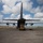 75th EAS Executes Tactical Airlift Mission in Manda Bay, Kenya A large military transport aircraft, consistent with a C-130 Hercules, is photographed from the rear on a tarmac or airstrip. A yellow forklift or ground support vehicle is positioned beneath the open rear cargo ramp, suggesting active loading or unloading operations. Several helicopters are visible in the background on the right side of the airfield.