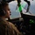 75th EAS Executes Tactical Airlift Mission in Manda Bay, Kenya A military aviator in multicam/OCP uniform is seated in an aircraft cockpit, viewed from a side profile angle. The pilot is wearing Bose aviation headphones with a boom microphone and is looking forward through the cockpit windscreen. A heads-up display (HUD) with green and purple reflections is visible in the foreground, along with various cockpit instruments.