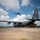 75th EAS Executes Tactical Airlift Mission in Manda Bay, Kenya A U.S. Air Force C-130 Hercules transport aircraft (tail number 83178, marked AMC) is parked on a tarmac with its propellers spinning, while a yellow front-end loader is positioned at its rear cargo ramp for loading or unloading operations. A military helicopter, likely a UH-60 Black Hawk, is visible in flight in the background. The scene appears to depict a forward operating base or austere airfield logistics operation.
