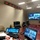 A civilian individual in a dark suit is seated at a conference room table participating in a multi-party video teleconference. The room appears to be a military or government command facility, equipped with multiple screens displaying remote participants, a digital clock display on the wall, and professional AV equipment. Several remote participants, including individuals in military uniforms, are visible on the large wall-mounted display screens.