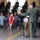 A military officer in a green flight suit is delivering a speech at a podium with a microphone inside what appears to be an aircraft hangar. The audience includes a mix of uniformed military personnel seated on bleachers in the background and civilians seated in chairs in the foreground. The setting and attire suggest a formal military ceremony, such as a change of command, promotion, or award presentation.