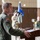 A military officer in a green flight suit is speaking at a podium during what appears to be a formal ceremony. He wears a distinctive patch on his left shoulder featuring a winged lion on a blue and orange background. The setting appears to be an indoor military facility with flags and other personnel visible in the background.
