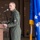 A military officer in an olive green flight suit is speaking at a podium with a microphone during what appears to be an official ceremony. He is wearing squadron patches on his uniform, including one that reads 'Pave Mafia,' and has lieutenant colonel oak leaf cluster insignia visible. The setting appears to be inside a military hangar with American and Air Force flags displayed in the background.