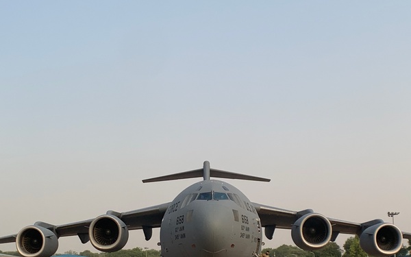 A large military transport aircraft, identifiable as a Boeing C-17 Globemaster III, is parked on a concrete tarmac at what appears to be a military airfield. The aircraft is photographed from a front-facing perspective, showing all four turbofan engines and its distinctive wide-body fuselage in gray livery. At least one ground crew member is visible near the forward entry door, suggesting pre-flight or post-flight servicing activities.