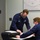 Two U.S. Coast Guard personnel in navy blue uniforms are reviewing documents at a desk in what appears to be an operations or briefing room. One standing individual, identified by name tape as 'BACEWICZ,' leans over to point at paperwork being reviewed by a seated colleague wearing glasses. Both individuals are wearing face masks, suggesting this was taken during the COVID-19 pandemic period.