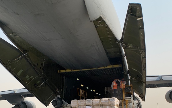A large cargo aircraft with its rear ramp open is being unloaded at an airport tarmac. A pallet of supplies marked with a USAID label is positioned on a cargo loader at the aircraft's rear door. Multiple ground personnel wearing high-visibility vests are overseeing the unloading operation.