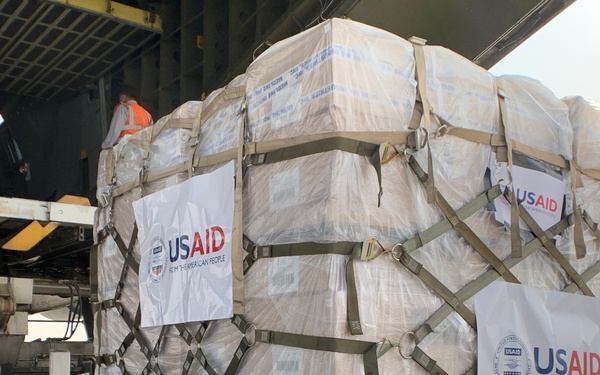 The image shows a large pallet of humanitarian aid supplies marked with USAID branding being loaded or unloaded from a military or cargo aircraft. The cargo is wrapped in clear plastic sheeting and secured with olive-drab cargo straps in a cross-hatch pattern. Multiple USAID labels reading 'FROM THE AMERICAN PEOPLE' are affixed to the exterior of the pallet.