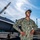 A U.S. Navy sailor named 'Brown' poses confidently on what appears to be a naval installation or aircraft carrier flight deck, standing in front of a military jet aircraft. The sailor is wearing the Navy Working Uniform (NWU Type III) in woodland digital camouflage pattern and is smiling broadly. A construction crane, American flag, and industrial buildings are visible in the background.