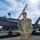 A U.S. Navy sailor in NWU Type I (digital camouflage) uniform poses smiling in front of a gray military jet aircraft, likely an F/A-18 Hornet or Super Hornet, on what appears to be a naval facility flight deck or tarmac. The sailor's name tape reads 'BROWN' and the branch tape reads 'U.S. NAVY'. Industrial cranes are visible in the background under a bright, sunny sky.