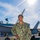 A U.S. Navy sailor named Brown, wearing NWU Type I (digital camouflage) uniform, poses smiling on what appears to be an aircraft carrier flight deck. A military jet aircraft (likely an F/A-18 Hornet or Super Hornet) is visible in the background along with a crane structure. The photo is taken in bright sunlight with lens flare visible.