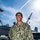 A U.S. Navy sailor named Brown, wearing NWU Type III (green digital camouflage) uniform, poses smiling on what appears to be the flight deck of an aircraft carrier. A military jet aircraft, likely an F/A-18 Hornet or Super Hornet, is visible in the background along with shipyard cranes, suggesting the vessel is in port or undergoing maintenance. The photo appears to be an official or semi-official portrait taken in bright daylight.