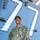 A U.S. Navy sailor in NWU Type III (green digital camouflage) uniform stands confidently aboard a naval vessel, smiling broadly at the camera. The sailor wears glasses, a green patrol cap, and has a name tape reading 'BROWN' with a 'U.S. NAVY' chest tape visible. The background shows the gray superstructure of a warship with the number '77' outlined in white lights.