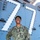 A U.S. Navy sailor named Brown is photographed smiling on the deck of a naval vessel. The sailor is wearing the standard Navy Working Uniform (NWU Type I) in digital camouflage pattern with 'U.S. NAVY' and 'BROWN' name tape visible. The background shows the gray superstructure of a warship with the number '77' illuminated in white lights.