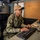 A U.S. Navy sailor wearing NWU Type III digital camouflage uniform is seated at a desk working on a computer keyboard. The sailor's name tape reads 'BROWN' and their cap displays 'USS GEORGE' with 'CVN' visible, indicating assignment to a Navy aircraft carrier. The setting appears to be an office or administrative workspace aboard a naval vessel.