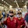 Three U.S. Navy personnel in red coveralls and black caps are engaged in what appears to be a briefing or instruction session inside a large hangar bay, likely aboard a naval vessel. The central figure, identified by name tape as 'CRUZHERNANDEZ,' is speaking and gesturing with a raised finger while wearing a black face mask. The setting and uniforms suggest a damage control or firefighting training context.