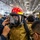 A person in full firefighting gear, including a red helmet with face shield, full-face respirator/SCBA mask, and tan/gold turnout coat, is being assisted by multiple people in donning or adjusting their equipment. The setting appears to be a large indoor facility, likely a hangar bay or industrial space, with numerous other personnel in dark uniforms visible in the background. This appears to be a firefighting training or drill exercise.