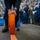 A military or coast guard training exercise is taking place in what appears to be a large indoor facility, likely a ship's hangar bay. A person in dark blue coveralls is unrolling an orange fire hose or similar emergency equipment along the deck floor while a group of uniformed personnel observe. The observers are wearing a mix of blue coveralls and camouflage uniforms, suggesting a multi-branch or training demonstration context.